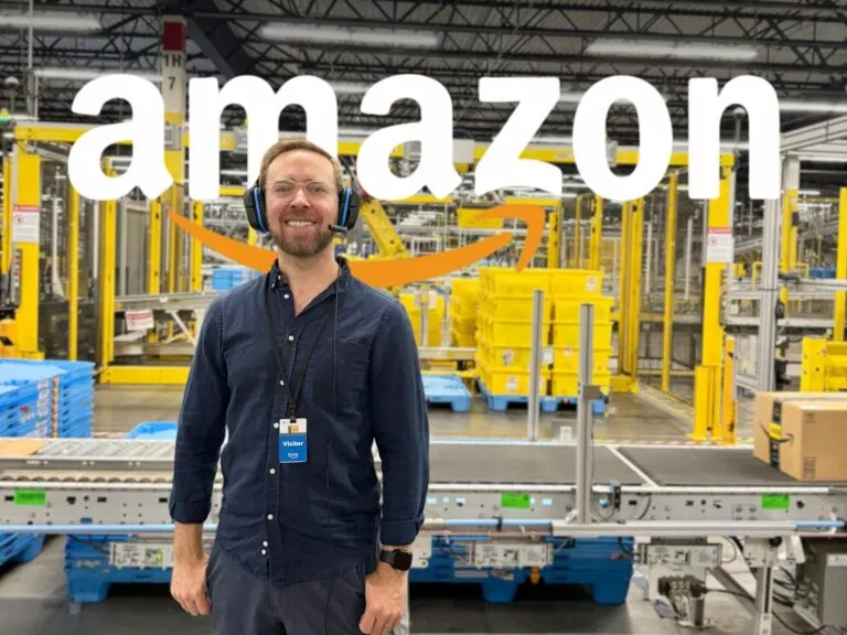Nick Gray in a headset and visitor badge, smiling in front of conveyor belts inside an Amazon warehouse